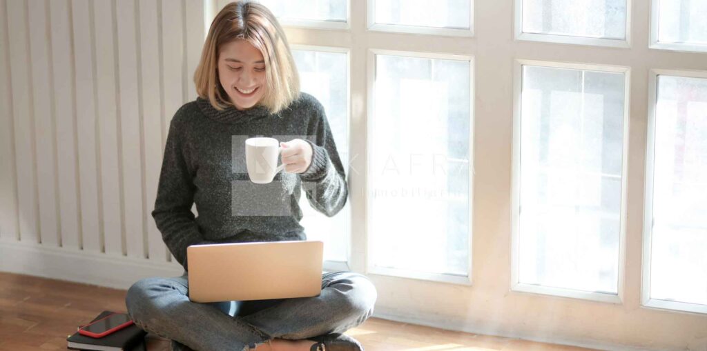 Mujer sentada en el suelo tomando café mientras está sosteniendo una laptop y la marca de agua de Inmobiliaria KIAFRA