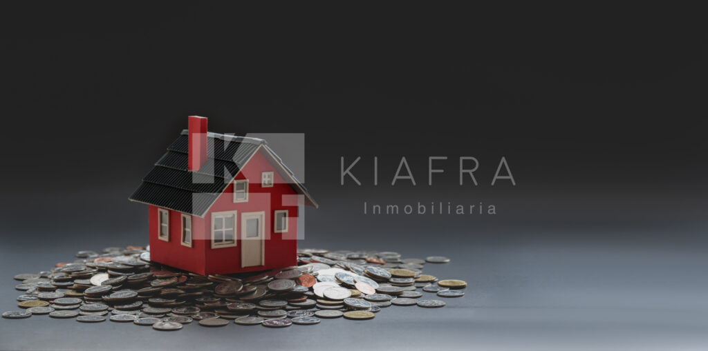 Casita de madera roja sobre una pila de monedas y la marca de agua de Inmobiliaria KIAFRA