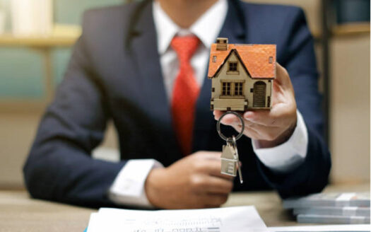 Mano de un asesor inmobiliario extendiendo una casita de madera con techo rojo y unas llaves con un llavero con forma de casita y el marco de Inmobiliaria KIAFRA