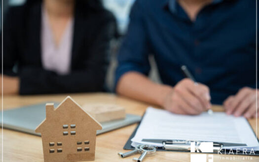 Casita de madera colocada en un escritorio y en el fondo se ve una pareja firmando un contrato de compraventa con el marco de Inmobiliaria KIAFRA