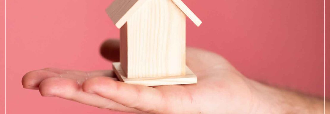Mano sosteniendo una casita de madera con el fondo rosa y el marco de Inmobiliaria KIAFRA